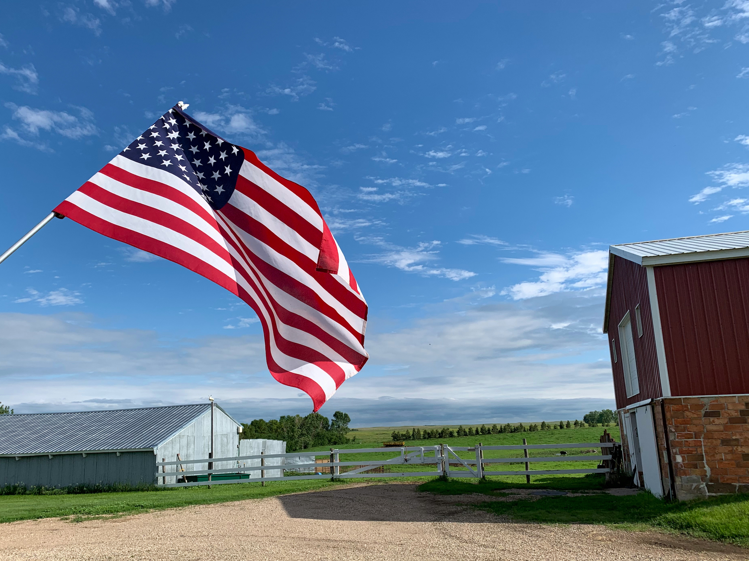ow angle close up view of American Flag against a blue sky with fluffy white clouds with american farm buildings in background