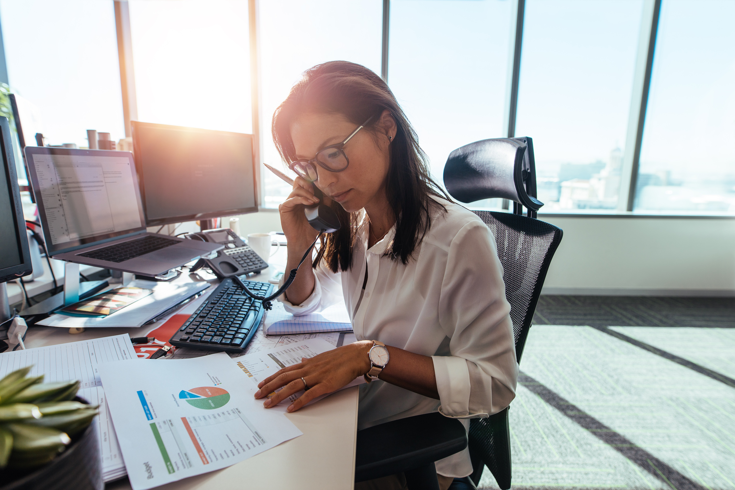 Woman entrepreneur looking at business papers while talking over phone. Woman sitting at her desk in office working with papers while seated in front of desktop computers.