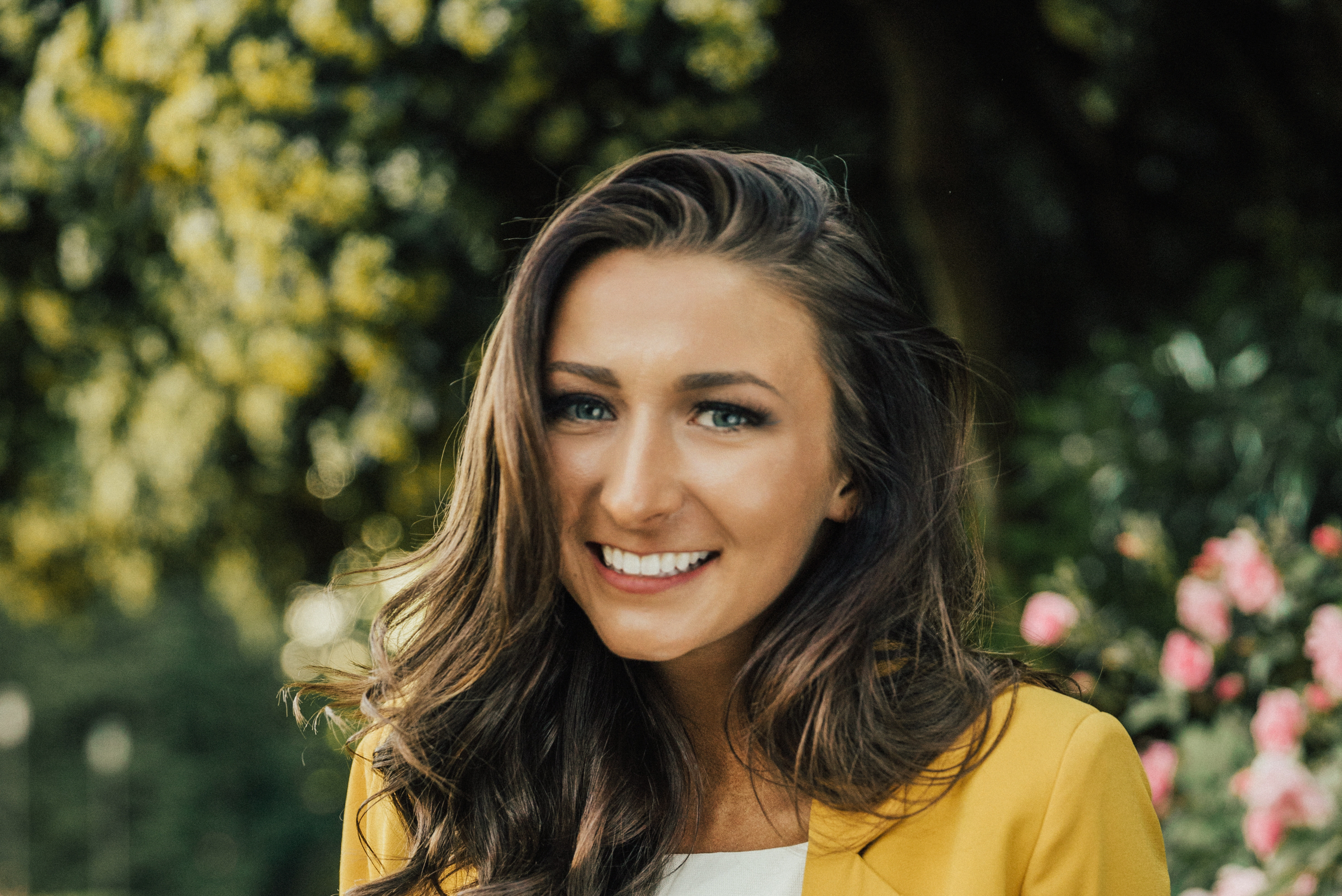 A woman with long brown hair and a yellow blazer stands in a garden.