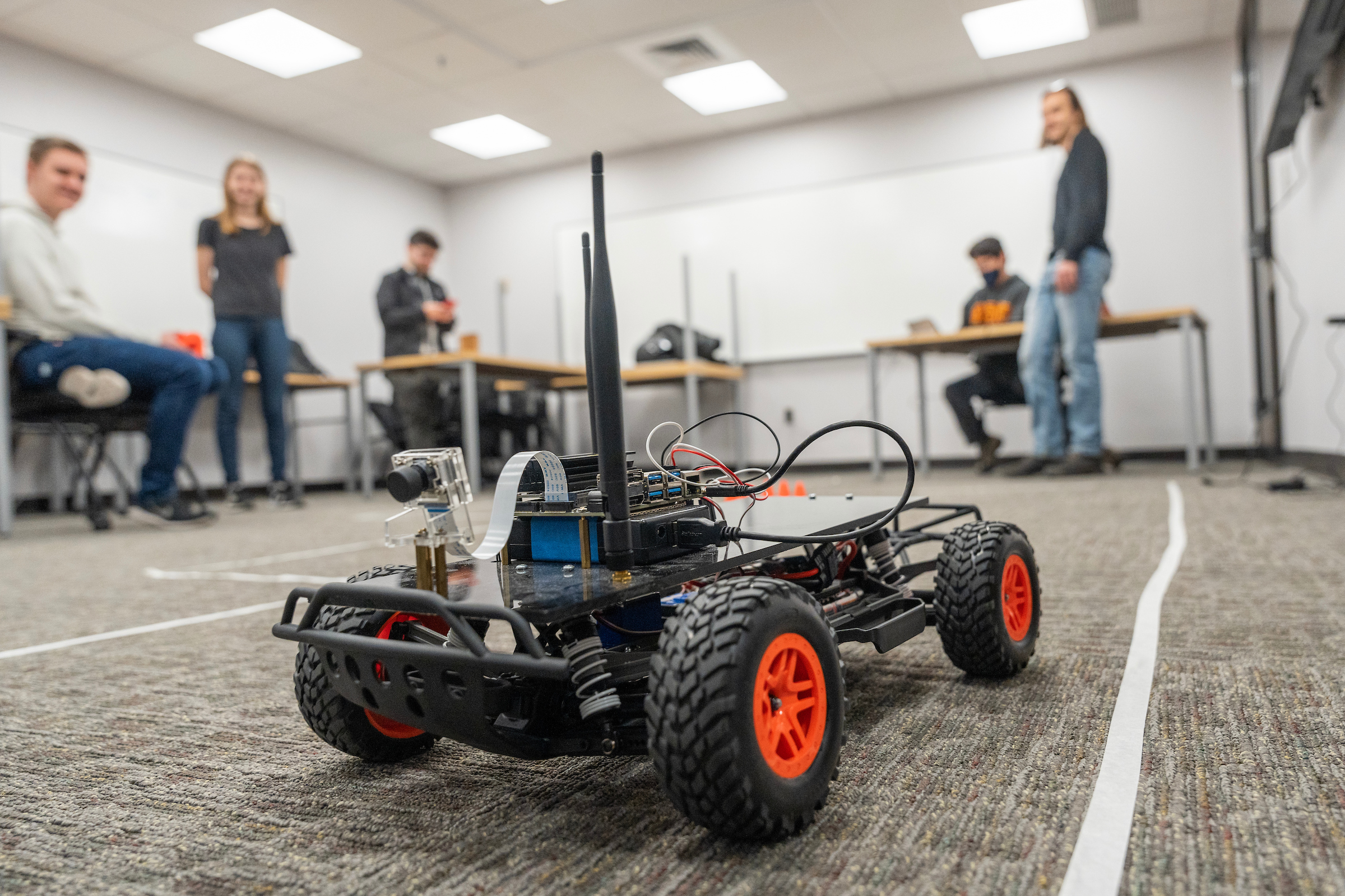 A small black robot on an obstacle course with four students and an instructor in the background.