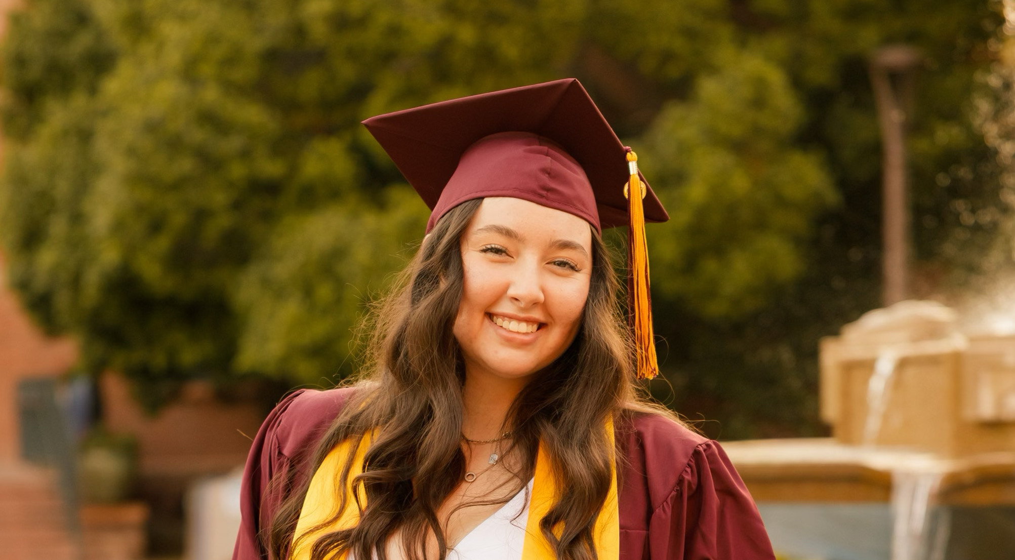 A woman with long brown hair wears a maroon graduation cap and gown.