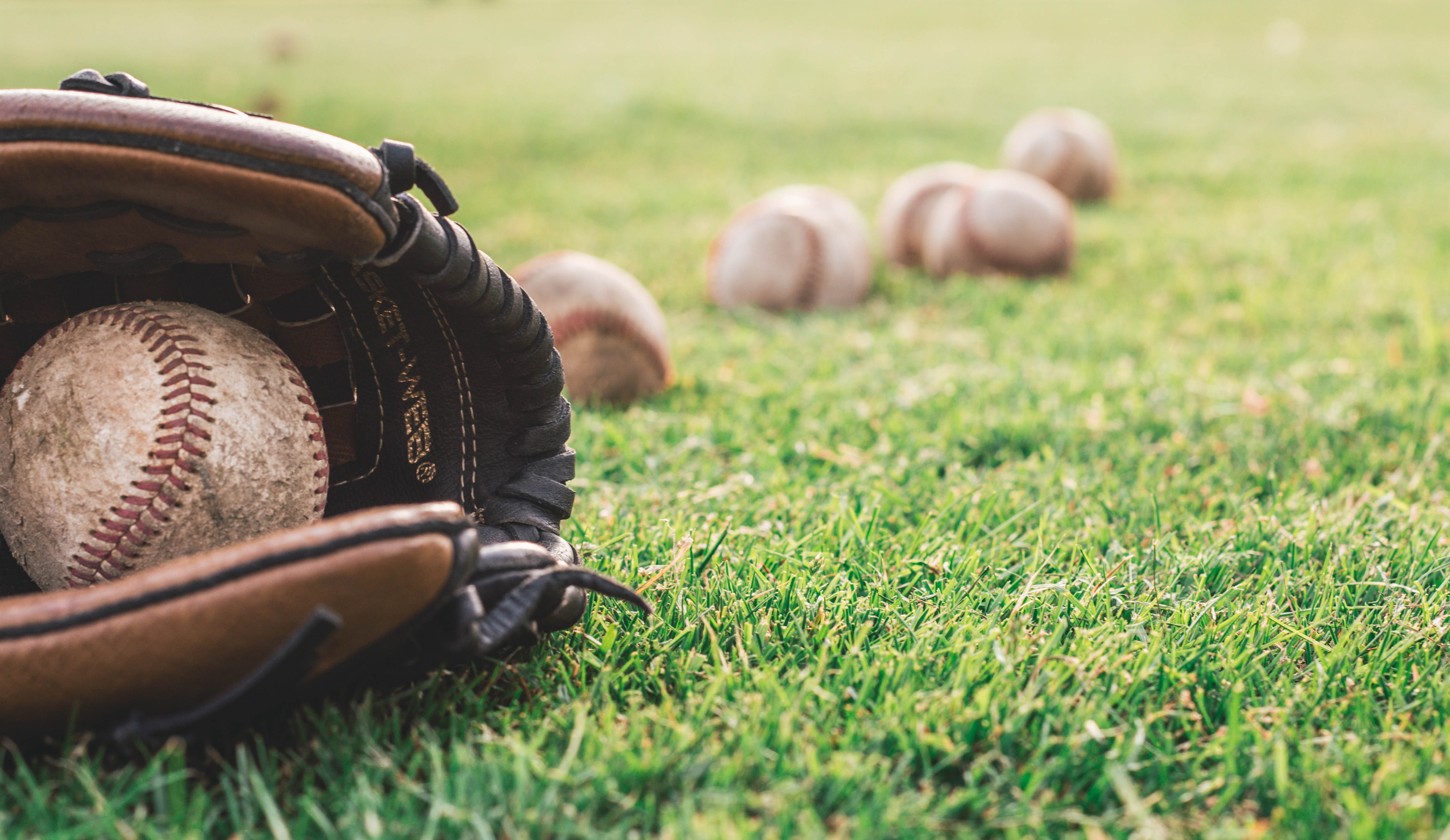 A baseball glove with several baseballs lying on green grass.