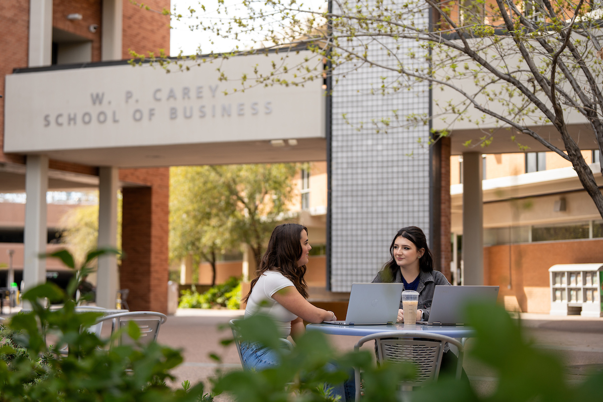 The image depicts two young women sitting at an outdoor table with laptops, engaged in conversation. They are located in front of the W. P. Carey School of Business building. The setting is relaxed, with greenery in the foreground and the business school's entrance visible in the background.